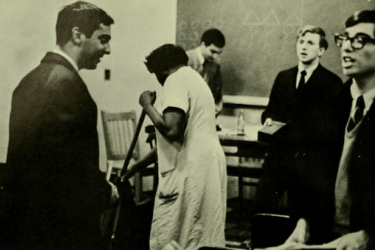 Black woman sweeping a classroom floor while white male students have a discussion