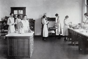 White female students in Home Economics class working in aprons at countertops with a Black woman at work in the background