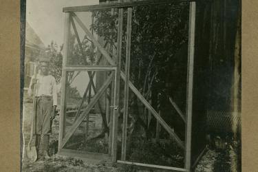 Black man with a shovel standing next to a tree surrounded by a wire breeding cage