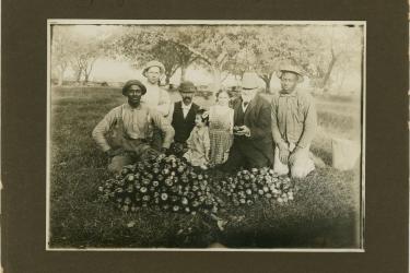 Two Black men kneel with a group of white men and children behind piles of sprayed apples