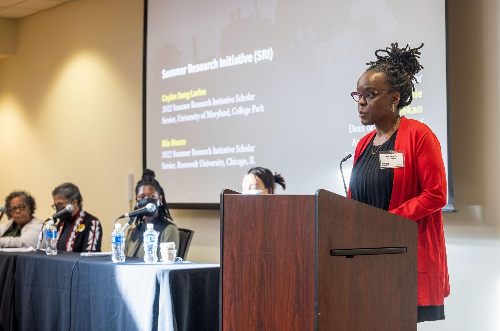 Dr. Stephanie Shonekan standing at a podium next to Session 2 panelists