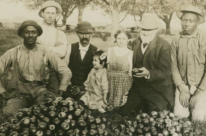 Two Black men kneel with a group of white men and children behind piles of sprayed apples.