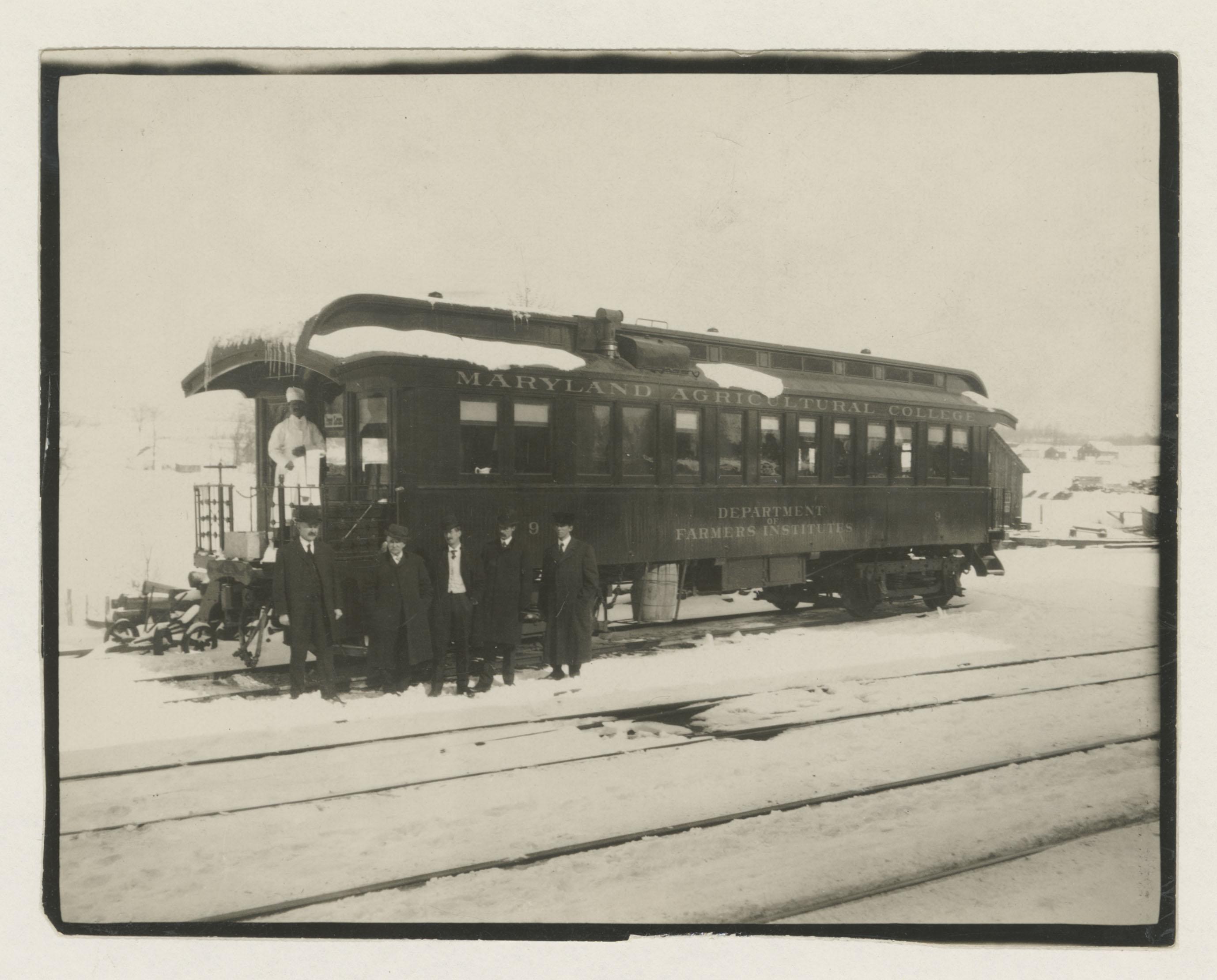 sepia toned photo of a train car with five white men in suits standing in front and a Black men in a chef's uniform behind them on the train